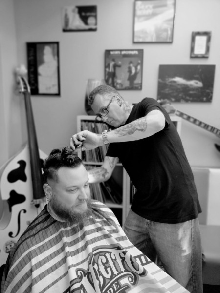 Raymon knipt een klant met baard in de zaak, met vintage posters en een contrabas